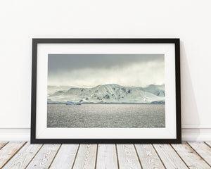 A blue iceberg floats before white Antarctic mountains beneath a low, clouded sky.