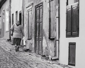 Romanian street photograph of a woman carrying bags past old shuttered buildings