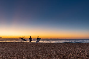 Three surfers stand and move across sand beside the ocean at dawn.